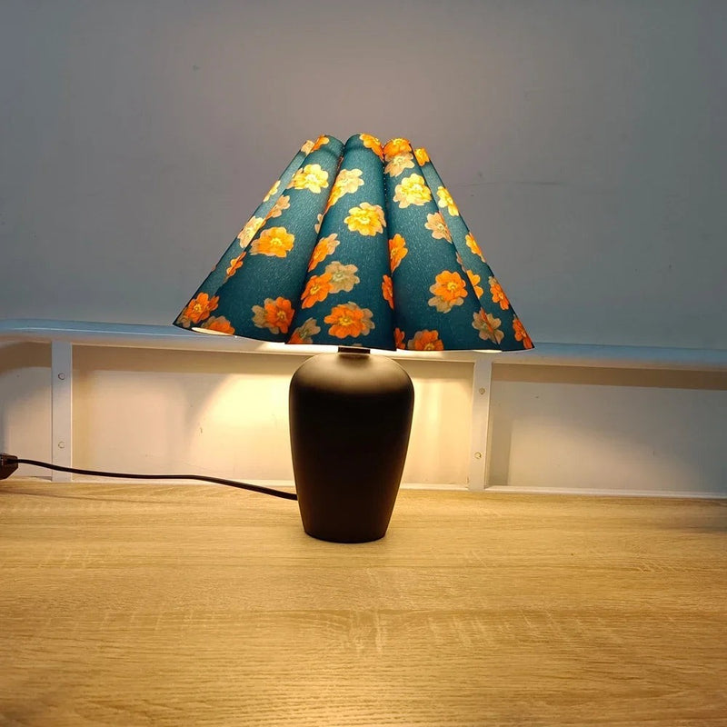 Lampe de table Dimoree avec base noire mate et abat-jour plissé bleu à motifs de fleurs orange sur une table en bois