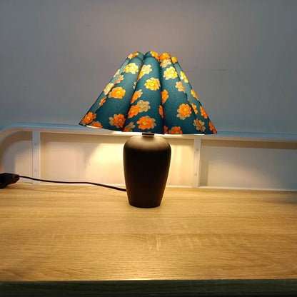Lampe de table Dimoree avec abat-jour plissé bleu à motifs floraux orange et pied en céramique noire sur bureau bois