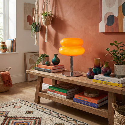 Lampe de table Dimoree en verre soufflé orange avec pied chromé sur un banc en bois dans un salon bohème.